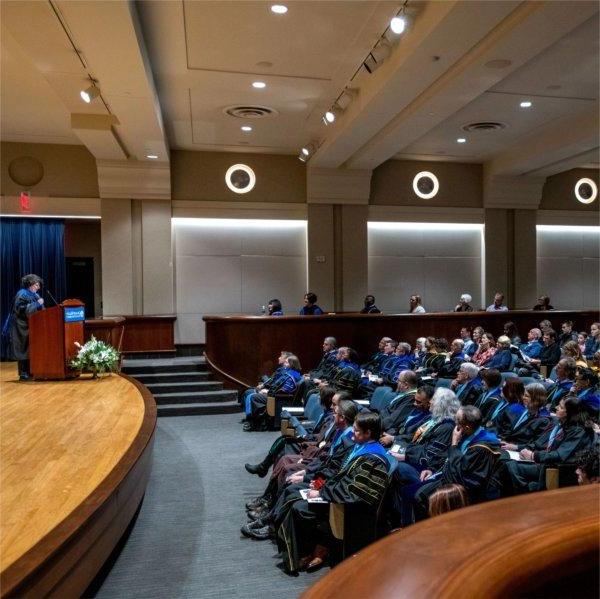 Samhita Rhodes, professor of engineering, speaks on stage in academic regalia to an audience at faculty convocation in Loosemore Auditorium